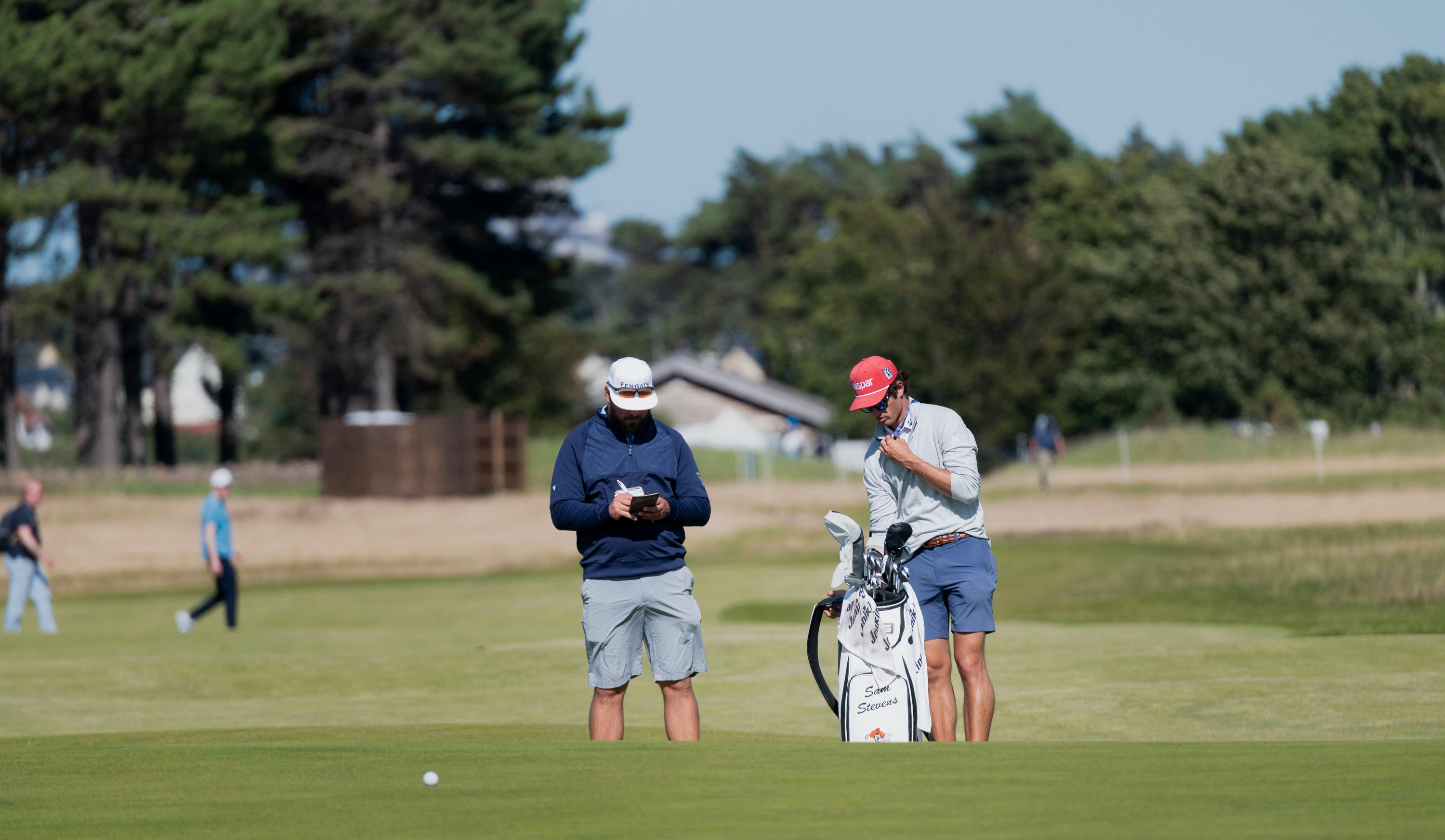 Two male golfers on a green, one writing on a notepad, the other looking at his golf bag.